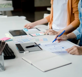 Web Design & Development Two people stand at a desk reviewing printed charts and notes, with a laptop, tablet, and office supplies nearby.